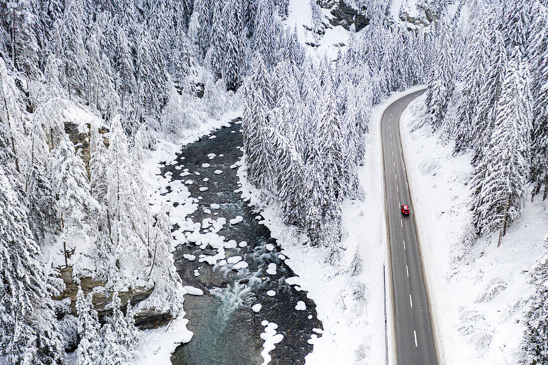 Auto auf der Bergstraße durch den verschneiten Wald und den gefrorenen Fluss, Luftaufnahme, Schweiz