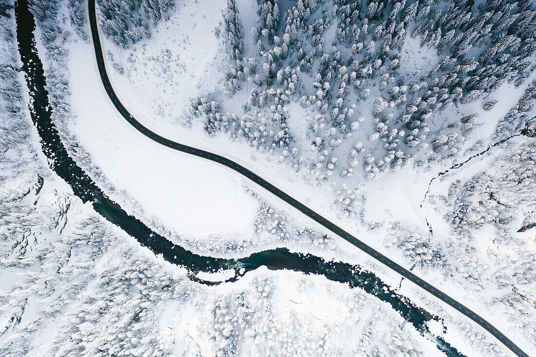 Luftaufnahme einer leeren Bergstraße, die neben einem gefrorenen Fluss im verschneiten Winterwald verläuft