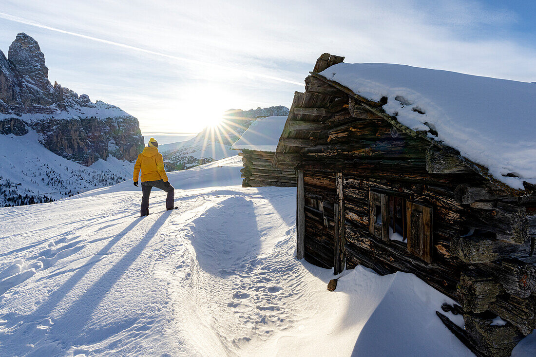 Rückansicht eines Mannes, der den Sonnenuntergang von einer traditionellen, mit Schnee bedeckten Holzhütte aus bewundert, Grödnerjoch, Dolomiten, Südtirol, Italien
