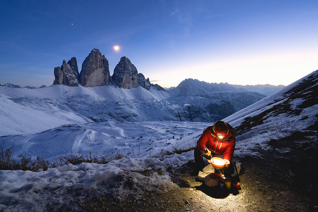 Wanderer bei der Zubereitung einer Mahlzeit mit einem Campingkocher und den Drei Zinnen im Hintergrund in der Abenddämmerung, Sextner Dolomiten, Südtirol, Italien