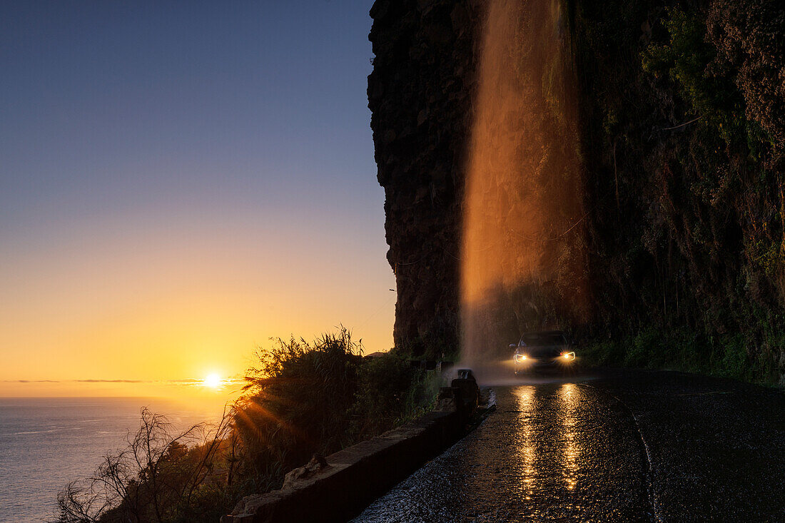 Car with on headlights passing under Anjos waterfall at dusk, Ponta do Sol, Madeira island, Portugal