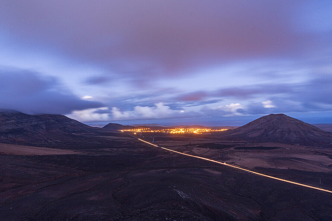 Lichter von Autospuren auf der Wüstenstraße am Fuße des Berges Tindaya in der Abenddämmerung, Aussichtspunkt Vallebron, Fuerteventura, Kanarische Inseln, Spanien