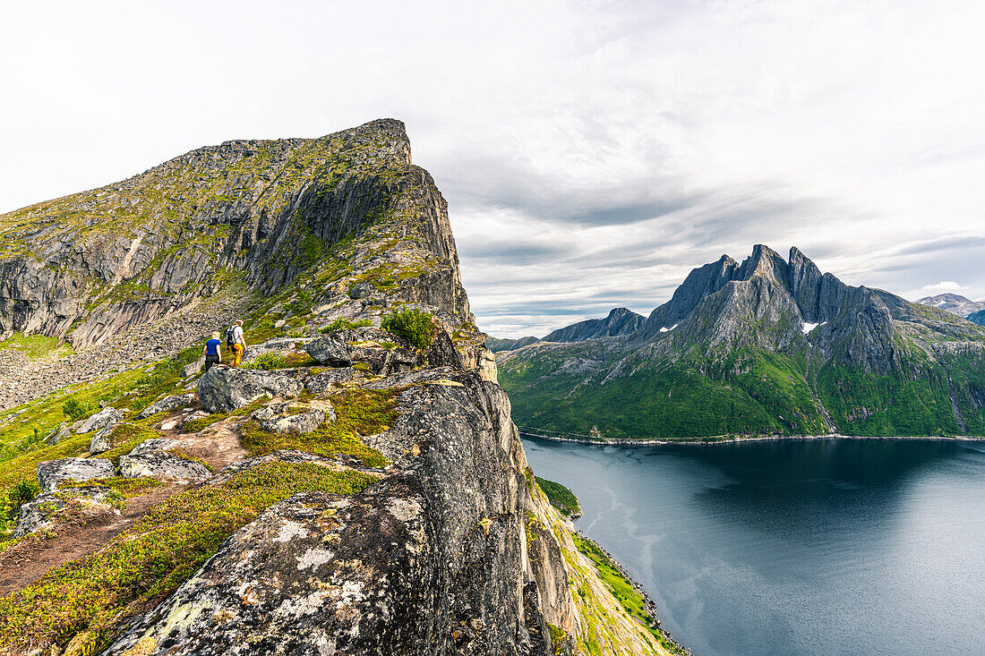 Zwei Personen wandern auf dem Grat zum Bardengipfel mit dem Mefjord und dem Berg Breidtinden im Hintergrund, Senja, Troms, Norwegen