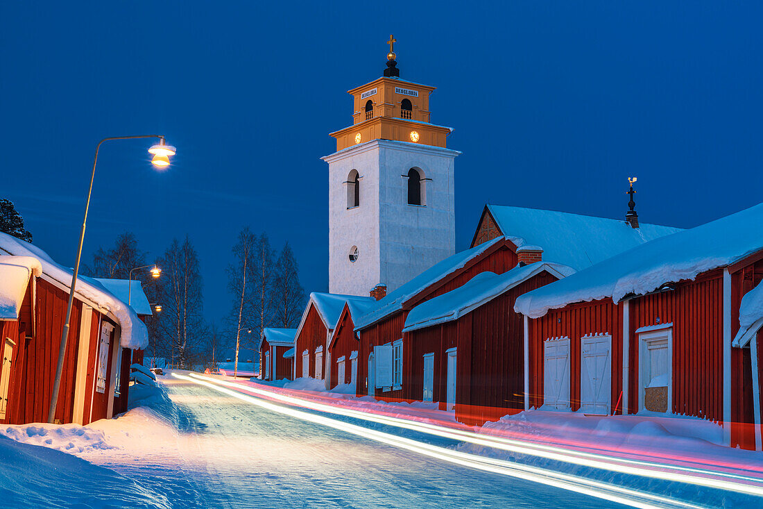 Lichterspuren von Autos auf der vereisten Straße, die durch die mittelalterliche, verschneite Kirchenstadt Gammelstad führt, Lulea, Schweden, bei Nacht