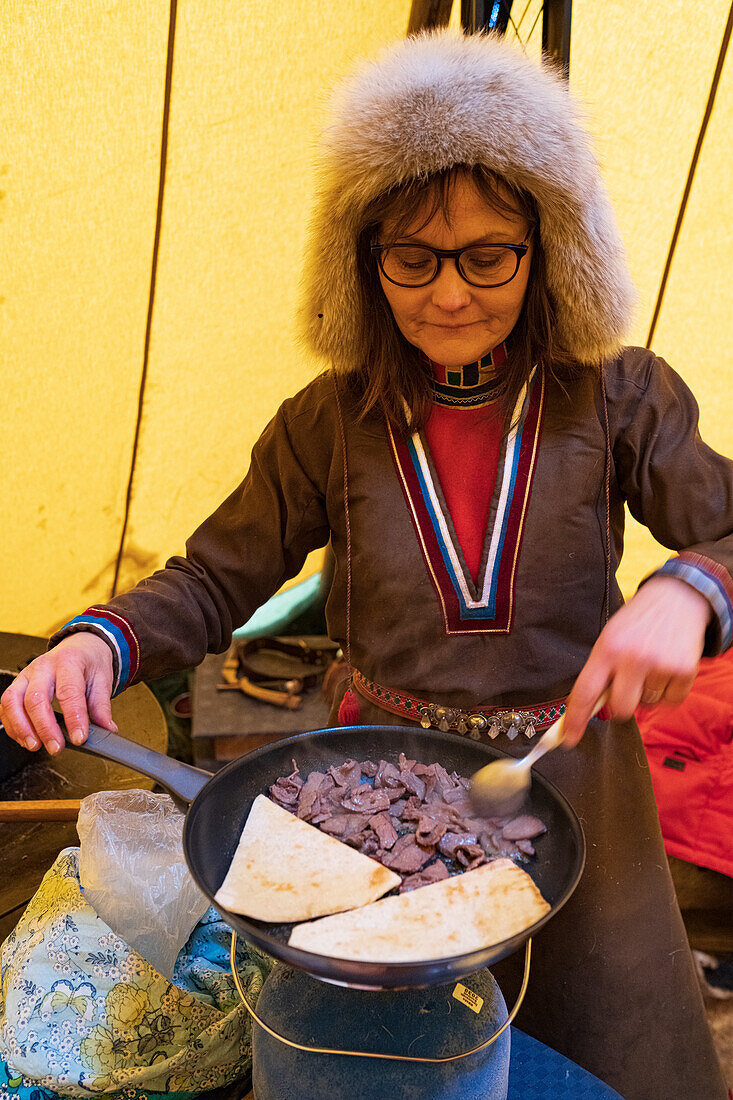 Porträt einer Frau in traditioneller samischer Kleidung beim Kochen von Rentierfleisch in einem Zelt, Lappland, Schweden