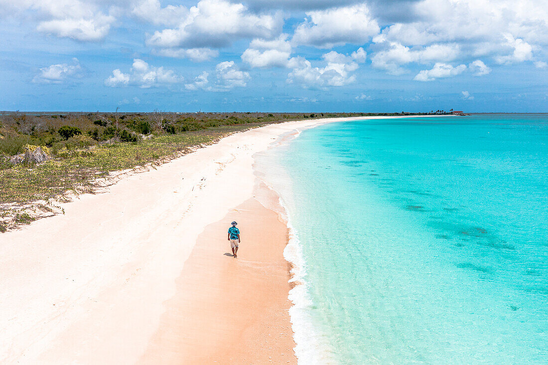 Luftaufnahme eines sorglosen Mannes mit Strohhut, der an einem idyllischen Strand spazieren geht, der vom türkisfarbenen Meer umspült wird, Antigua und Barbuda, Karibik