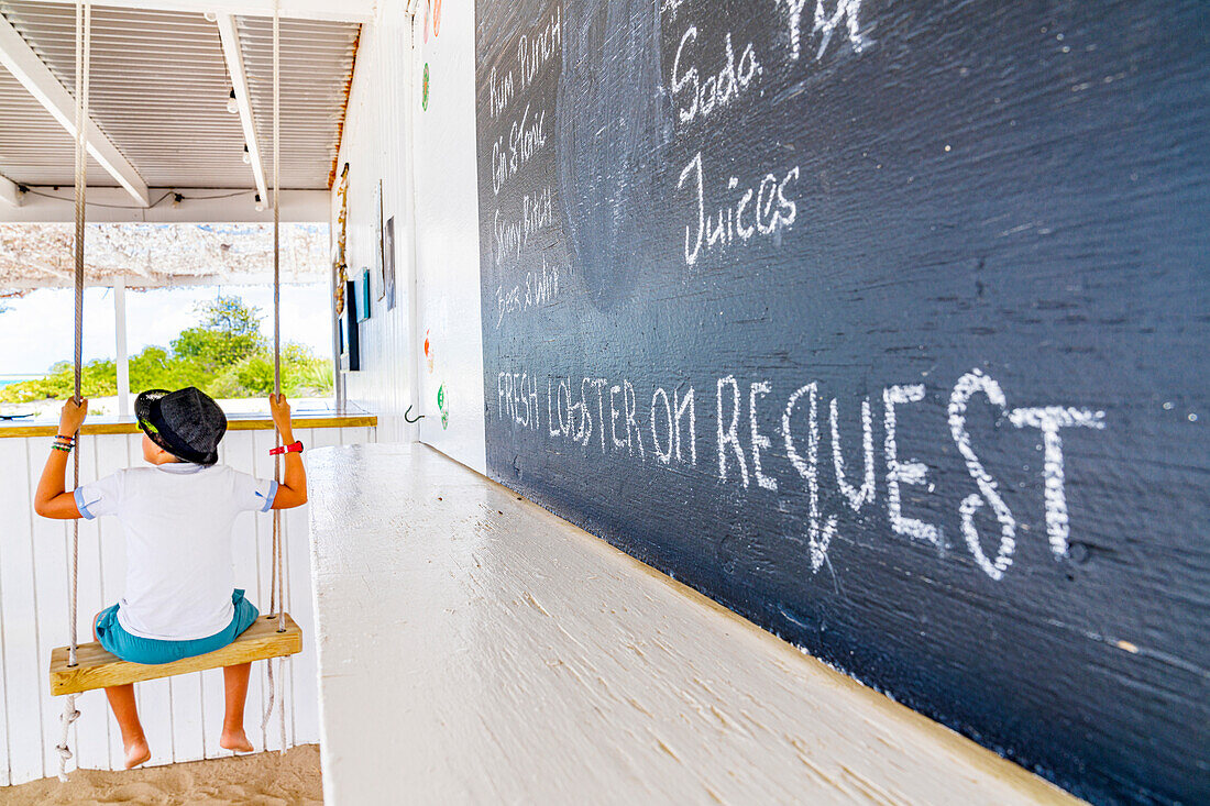 Happy little boy sitting on a swing in a beach bar in front of Caribbean Sea, Barbuda, Antigua & Barbuda, West Indies