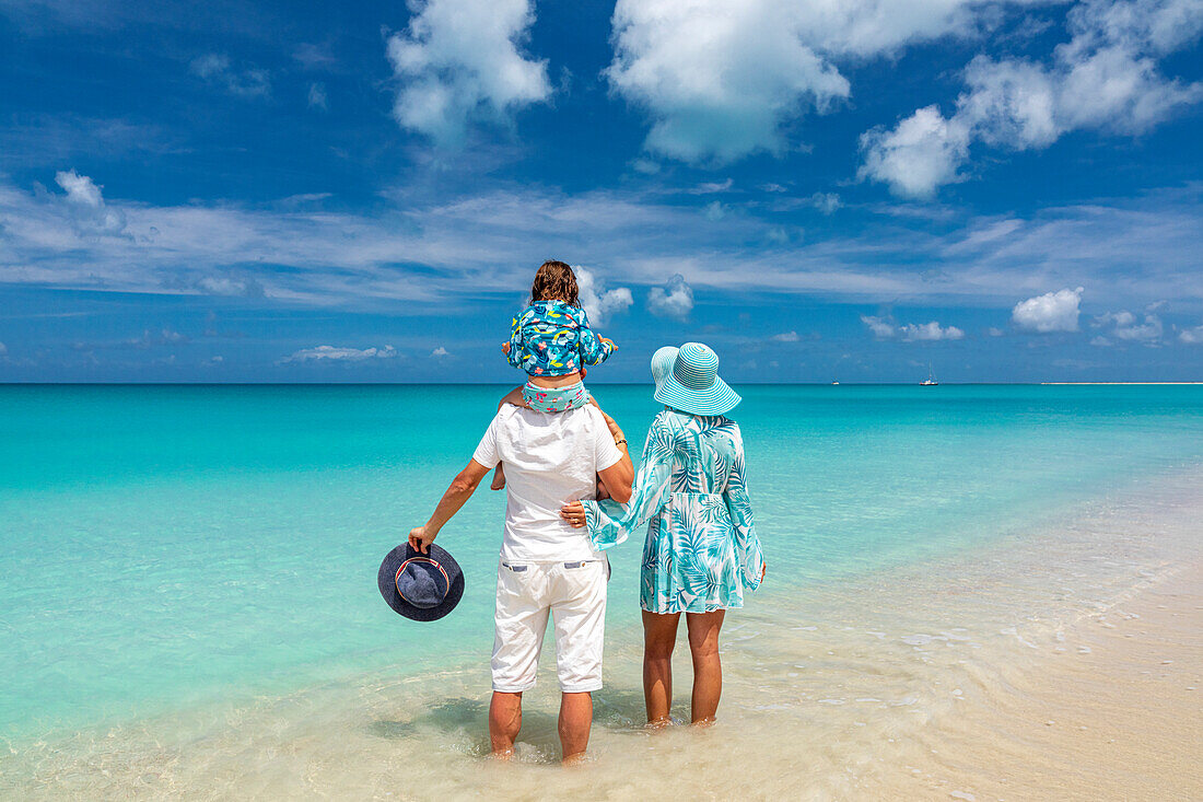 Glückliche Eltern stehen im klaren Wasser des Karibischen Meeres und tragen ein kleines Mädchen Huckepack, Antigua und Barbuda, Karibik
