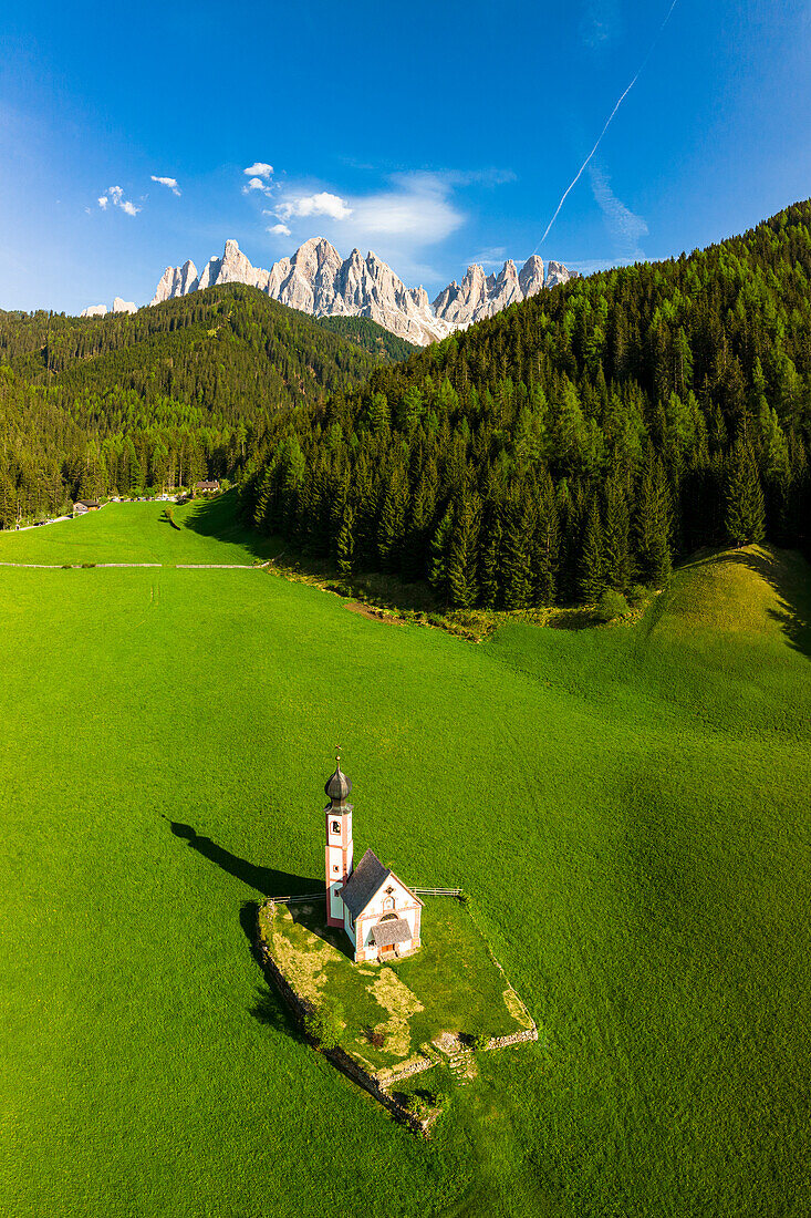 Die ikonische Kirche von Ranui und die Geislergruppe umgeben von grünen Wäldern im Frühling, Blick von oben, Funes-Tal, Südtirol, Italien
