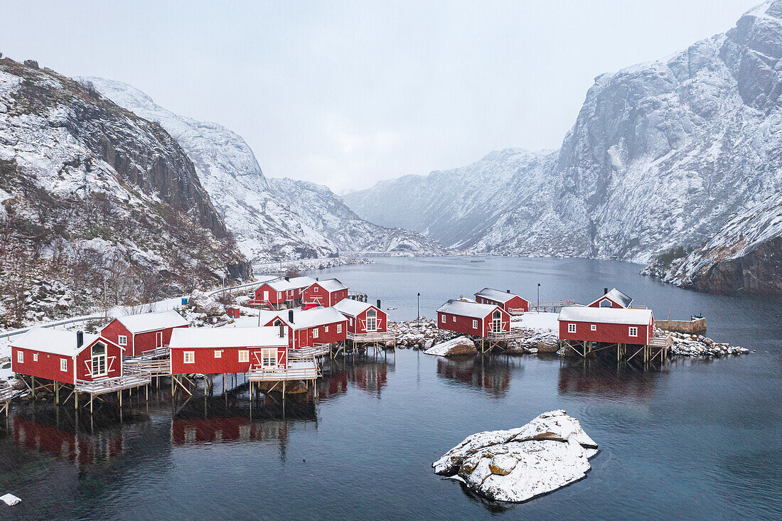 Nebliger Himmel über schneebedeckten Bergen und roten Rorbu-Hütten am Fjord, Nusfjord, Nordland, Lofoten, Norwegen