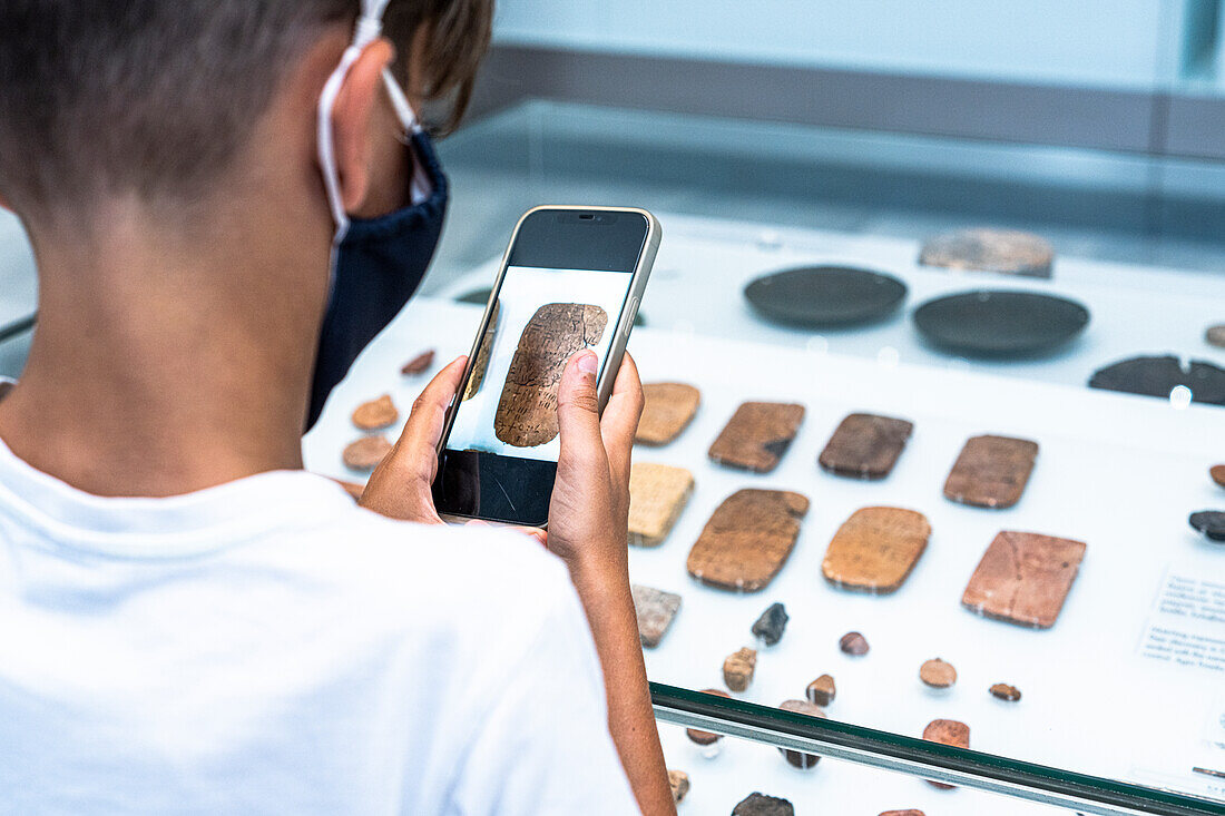 Little boy with face mask photographing old artcraft with smartphone, Heraklion Archaeological Museum, Crete island, Greece