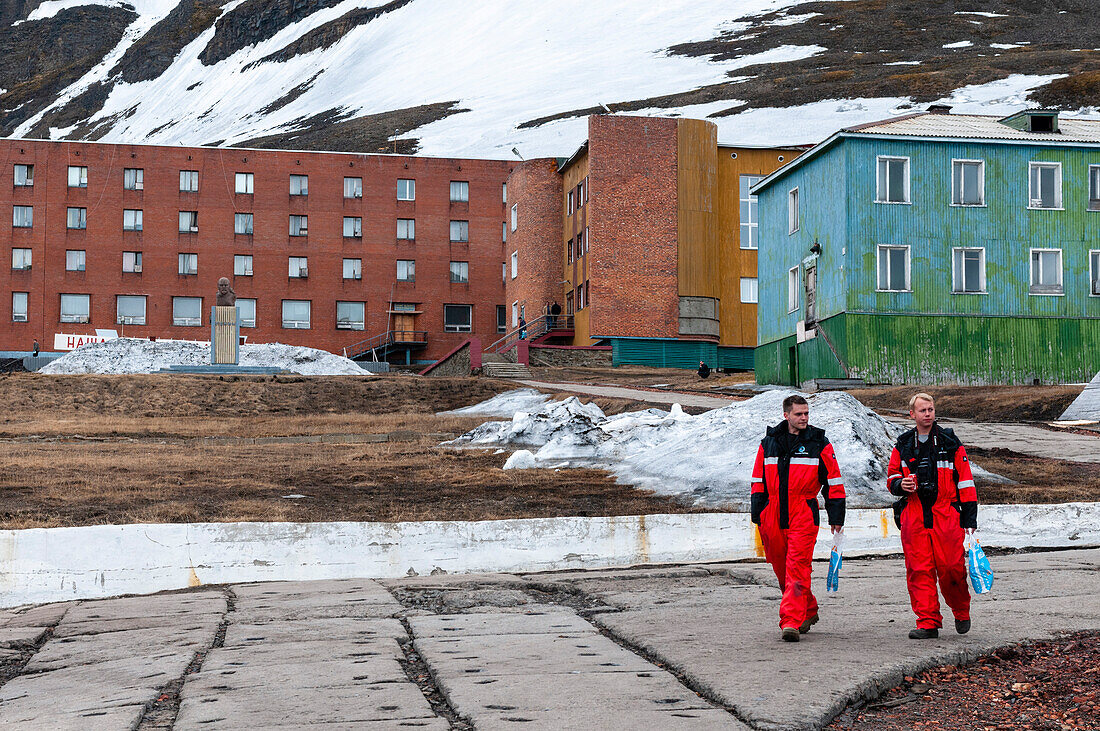 Männer in arktischer Schutzkleidung gehen an Wohnhäusern vorbei. Barentsburg, Insel Spitzbergen, Svalbard, Norwegen.