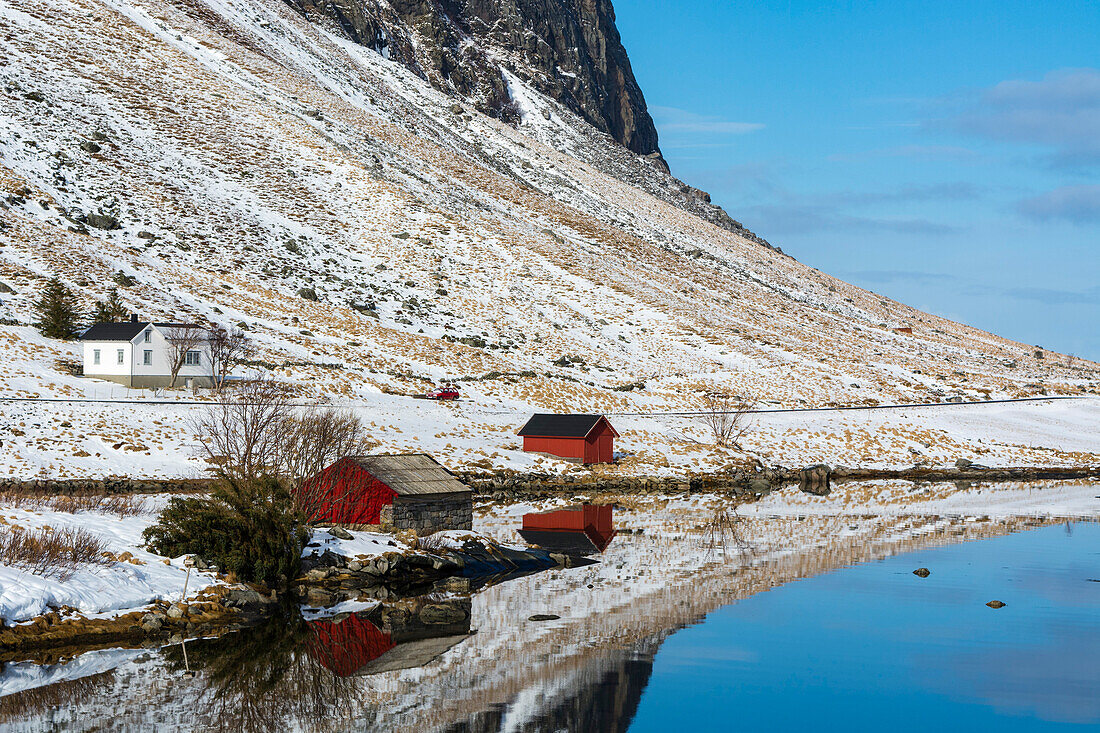 Rote Schuppen am Ufer, mit einem Haus weiter oben auf einem verschneiten Hügel. Eggum, Lofoten-Inseln, Nordland, Norwegen.