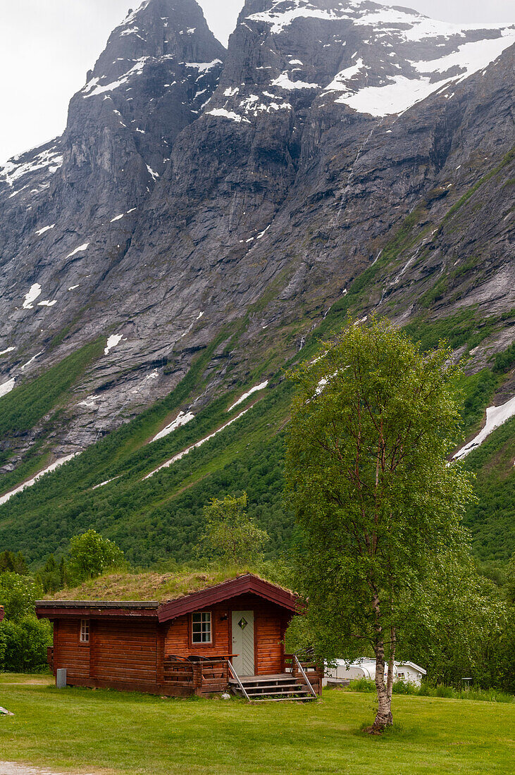 A primitive wood hut nestled in a verdant valley at the base of ice covered sheer cliffs. Trollstigen, Rauma, Norway.