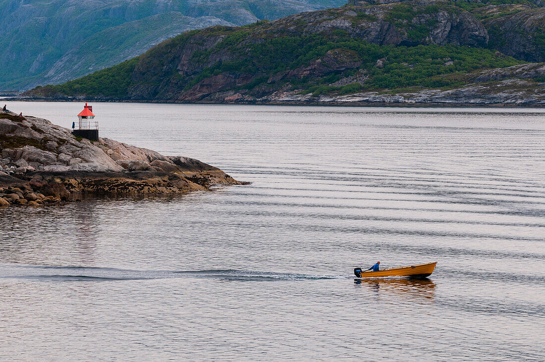 Ein kleines Motorboot durchquert die von Bergen gesäumten Gletschergewässer des Svesfjorden in der Nähe eines kleinen Leuchtturms. Svesfjorden, Norwegen.