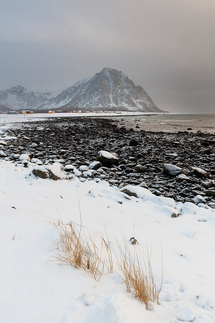 Eine schneebedeckte Küstenlinie mit einem felsigen Strand, Bergen und einem Dorf. Noss, Vesteralen-Inseln, Nordland, Norwegen.
