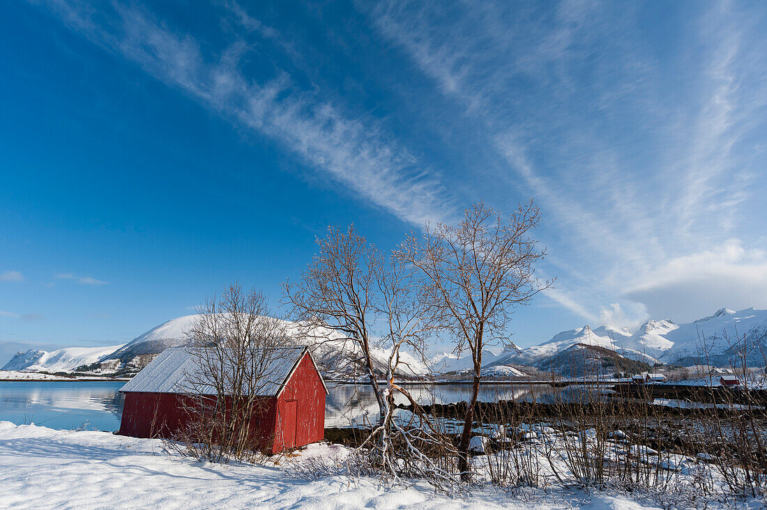 Eine rote Scheune oder ein Schuppen am Meeresufer. Schneebedeckte Berge in der Ferne. Vagan, Lofoten-Inseln, Nordland, Norwegen.