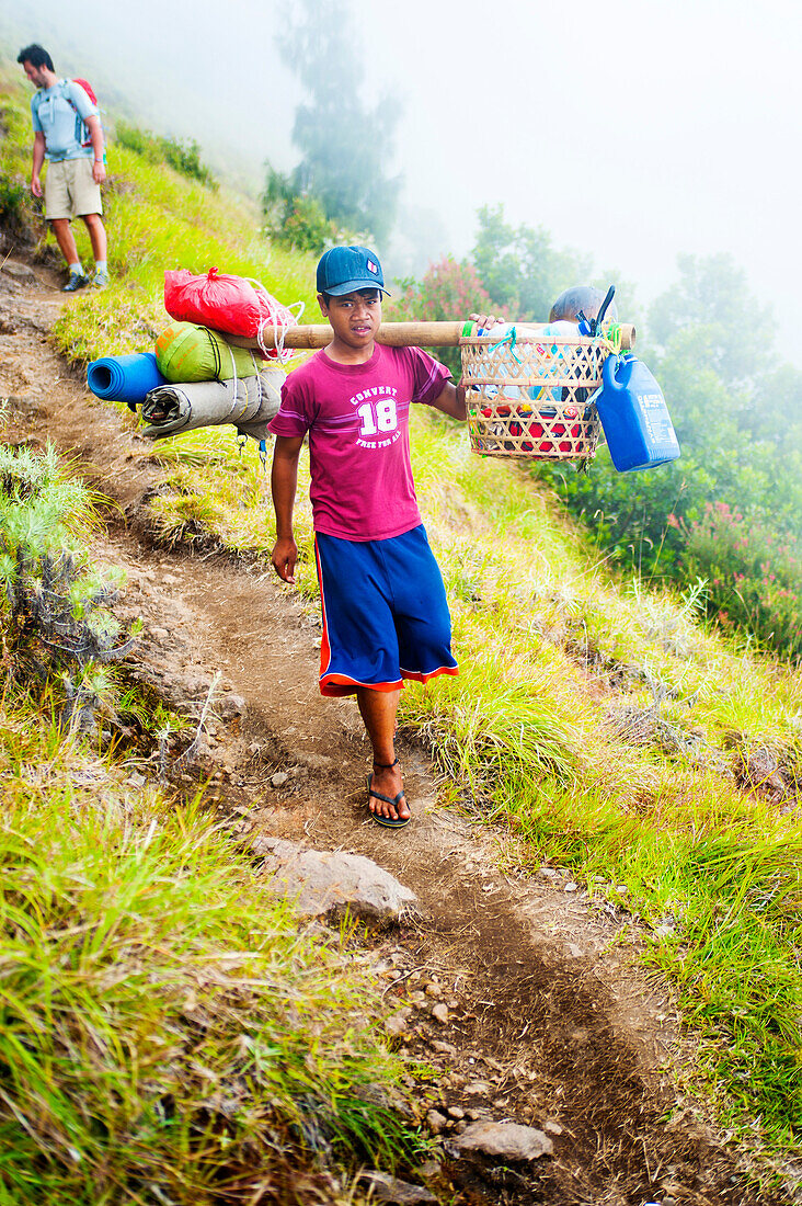 Junger Träger trägt Lebensmittel und Campingausrüstung auf dem Trek auf den Mount Rinjani, Lombok, Indonesien. Diese Träger sind unglaublich. Sie tragen die gesamte Verpflegung und Campingausrüstung für die dreitägige Wanderung zum Gipfel des Mount Rinjani, die etwa 35 kg wiegt, und sind dabei noch vor den Touristen unterwegs! Der Mount Rinjani (Gunung Rinjani auf Indonesisch) ist ein aktiver Vulkan auf der Insel Lombok, Indonesien. Mit einer Höhe von 3726 m ist er der zweithöchste Vulkan Indonesiens und bietet eine beeindruckende Aussicht über die gesamte Insel Lombok, im Westen über den Vulk