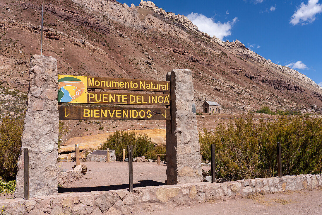 Schild für das Naturdenkmal Puente del Inca in den argentinischen Anden. Dahinter befindet sich die Kapelle Unserer Lieben Frau vom Schnee.