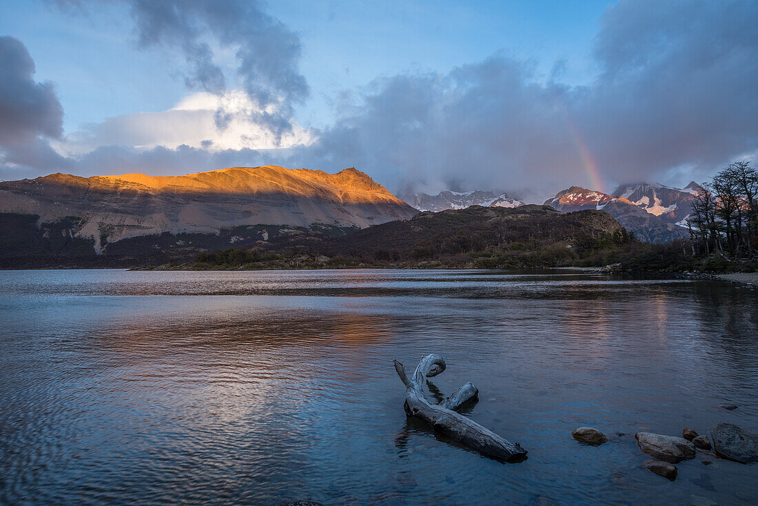 Der Sonnenaufgang beleuchtet die Loma de las Pizarras am Lago Capri im Nationalpark Los Glaciares bei El Chalten, Argentinien. Eine UNESCO-Welterbestätte in der südamerikanischen Region Patagonien. Das Fitz-Roy-Massiv wird rechts hinter dem Regenbogen von den Wolken verdeckt.