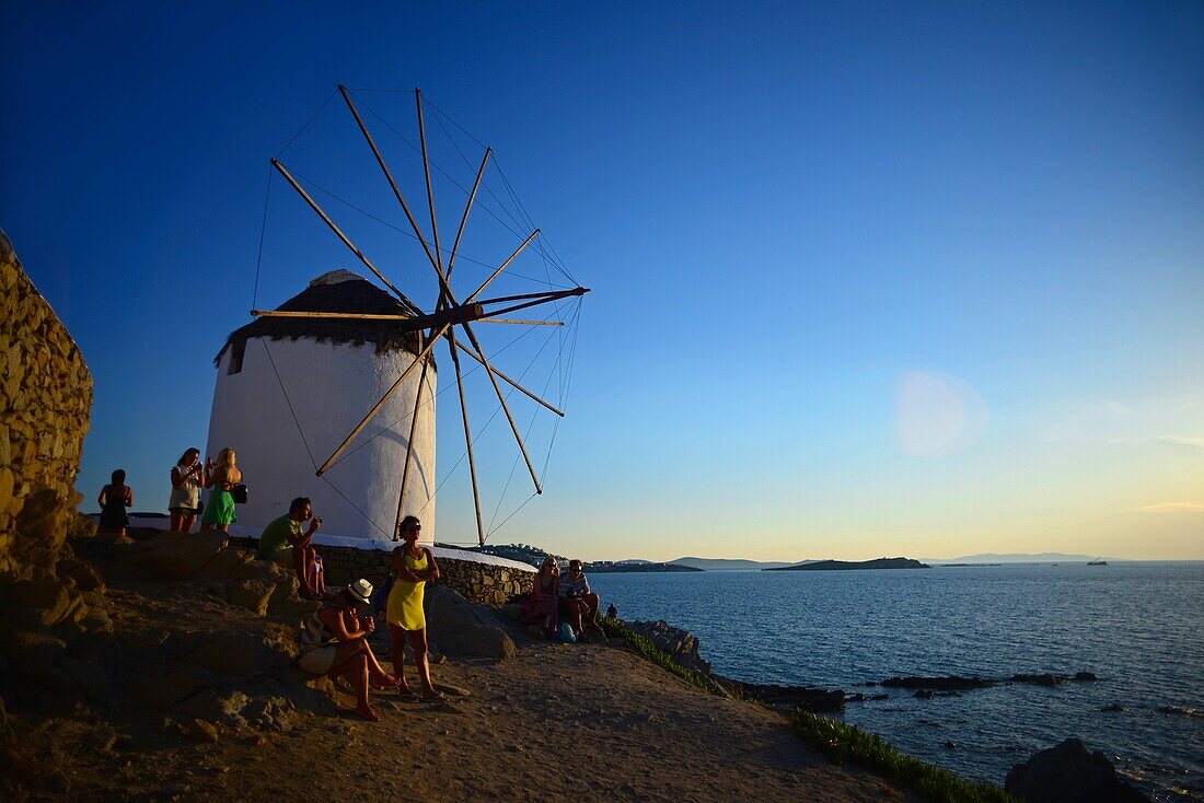 Menschen genießen den Sonnenuntergang vor traditionellen Windmühlen (Kato Milli) in Mykonos-Stadt, Griechenland