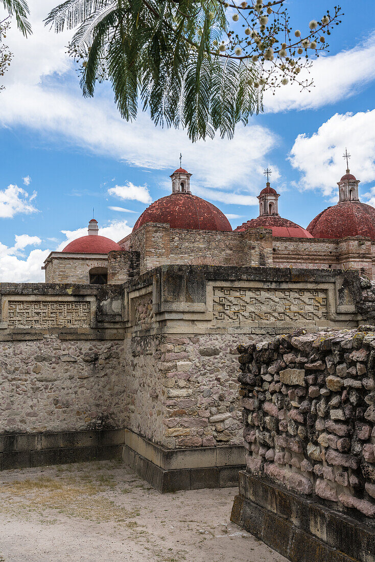 Laubsägearbeitstafeln aus Stein in den Ruinen der zapotekischen Stadt Mitla. Im Hintergrund sind die Kuppeln der Kirche von San Pablo zu sehen. Mitla, Oaxaca, Mexiko. Eine UNESCO-Welterbestätte.