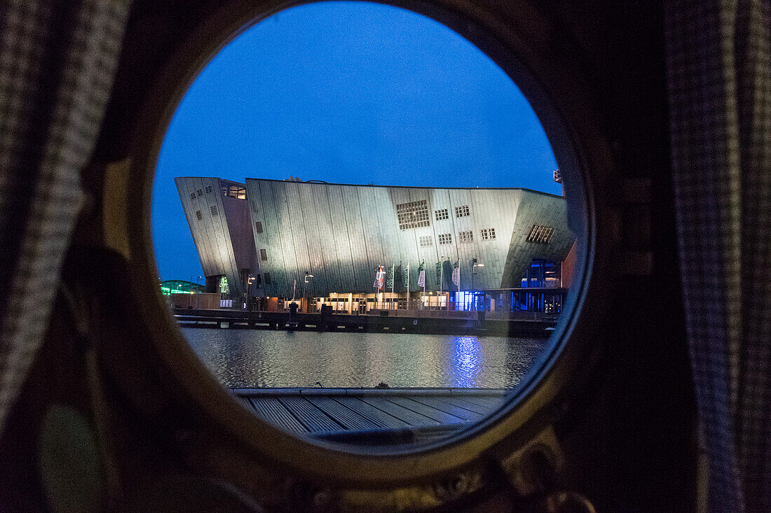 Amsterdam, Niederlande, Blick auf das Nemo Science Museum am Wasser durch das Bullauge eines Bootes bei Nacht.