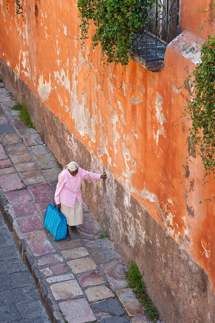 Ältere Frau geht auf der Straße und stützt sich mit der Hand an einer Wand ab; San Miguel de Allende, Guanajuato, Mexiko.