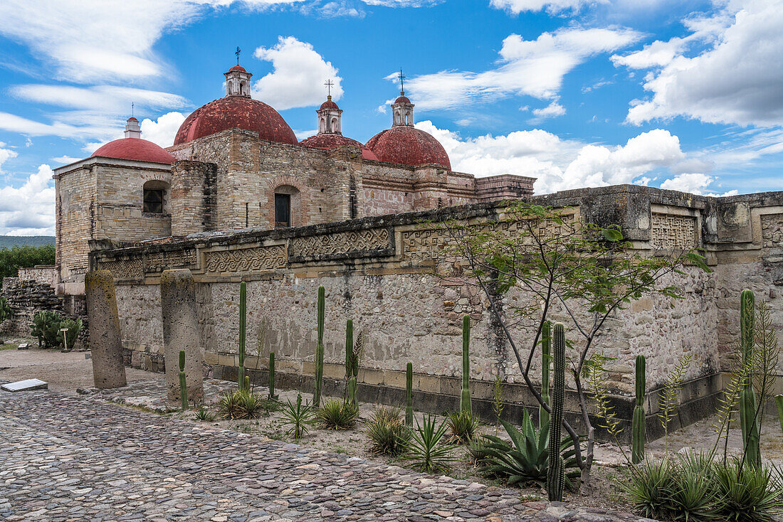 Kakteen, Steinsäulen und Laubsägearbeiten in den Ruinen der zapotekischen Stadt Mitla. Im Hintergrund ist die Kirche San Pablo zu sehen. Mitla, Oaxaca, Mexiko. Eine UNESCO-Welterbestätte.