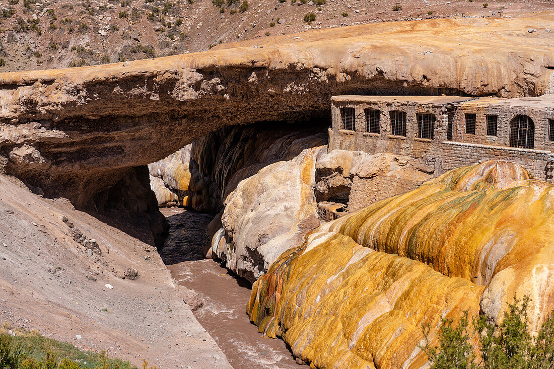 Farbenfrohe Travertinablagerungen der Mineralquelle in Puente del Inca in den argentinischen Anden, mit den Ruinen eines ehemaligen Kurortes.