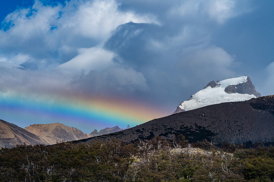 Ein Regenbogen neben dem schneebedeckten Cerro Solo im Los-Glaciares-Nationalpark in der Nähe von El Chalten, Argentinien, einem UNESCO-Weltkulturerbe. Im Vordergrund ist ein Wald aus Lenga oder Südbuchen zu sehen.