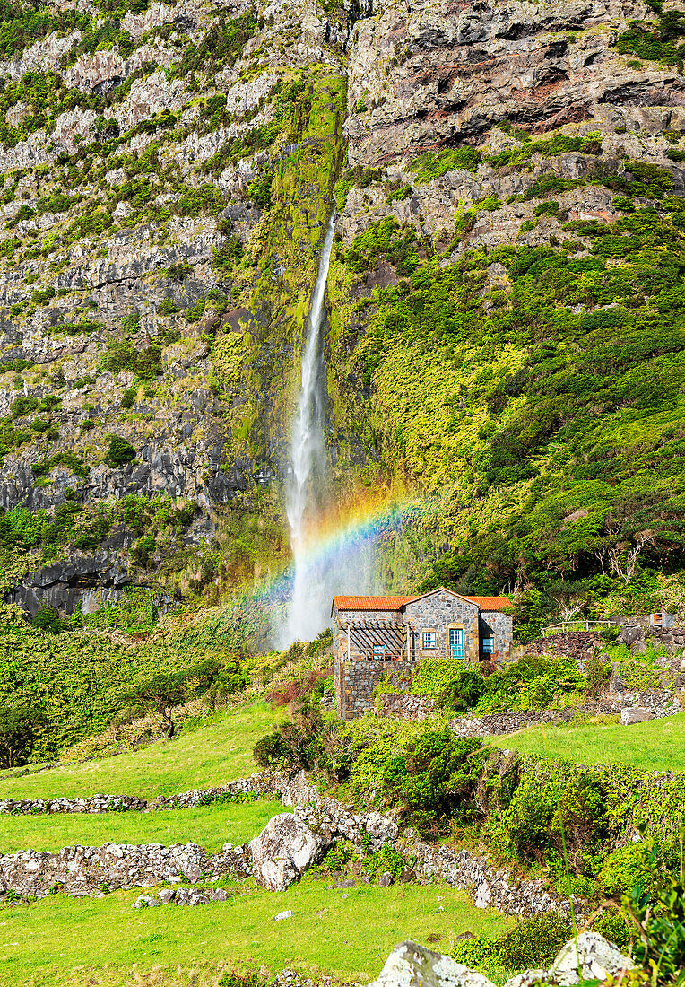 Blick auf den Wasserfall Poco do Bacalhau, der hinter einem Haus mit Regenbogen herunterfällt, Faja Grande, Lajes das Flores, Insel Flores (Ilha das Flores), Azoren-Archipel, Portugal, Atlantik, Europa
