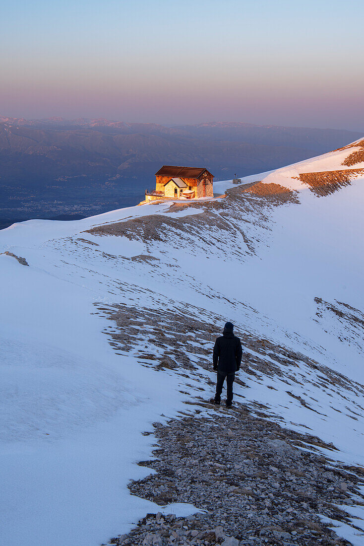 Winter view of a hiker admiring the sunset at Duca degli Abruzzi mountain hut, Campo Imperatore, Apennines, L'Aquila district, Abruzzo, Italy, Europe