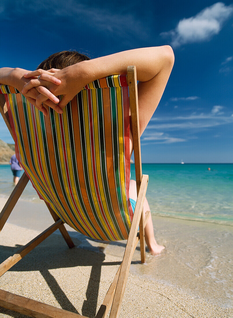 Rückansicht einer Frau, die sich in einem Liegestuhl am Meer am Strand von Porthcurno entspannt.