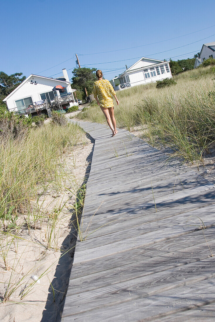Frau im Kaftan, die auf einem Holzsteg am Strand in Richtung Häuser geht