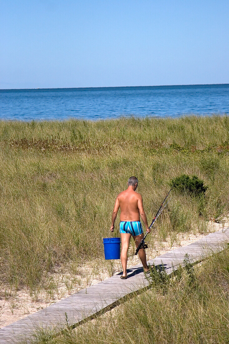 Älterer Mann geht auf einem hölzernen Strandweg und trägt eine Angelrute