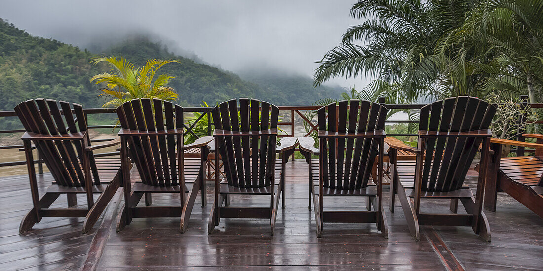 Sechs hölzerne Adirondack-Stühle sitzen in einer Reihe auf einem Holzdeck mit Blick auf die tief hängenden Wolken über einer bergigen Landschaft; Provinz Oudomxay, Laos.