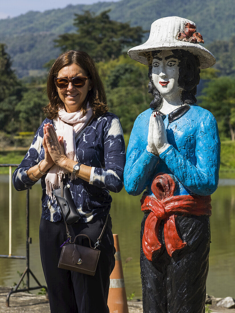 A Female Tourist Poses In A Similar Pose To A Thai Statue She Is Standing Beside; Tambon Pa Tueng, Chang Wat Chiang Rai, Thailand