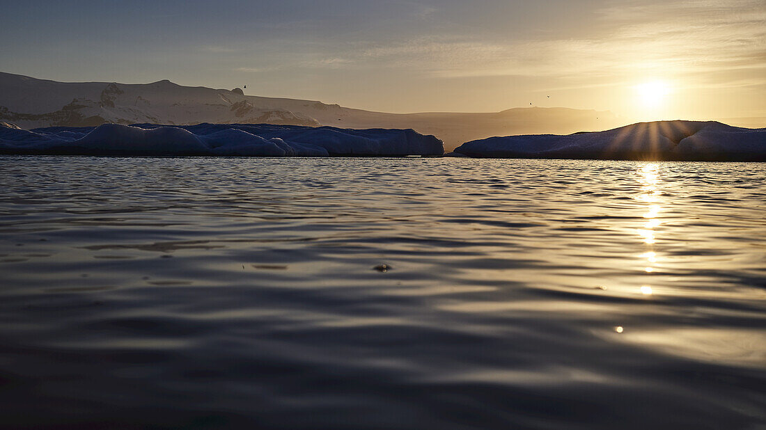 Goldener Sonnenuntergang über den Silhouetten der Berge entlang der ruhigen Wasser- und Küstenlinie; Island