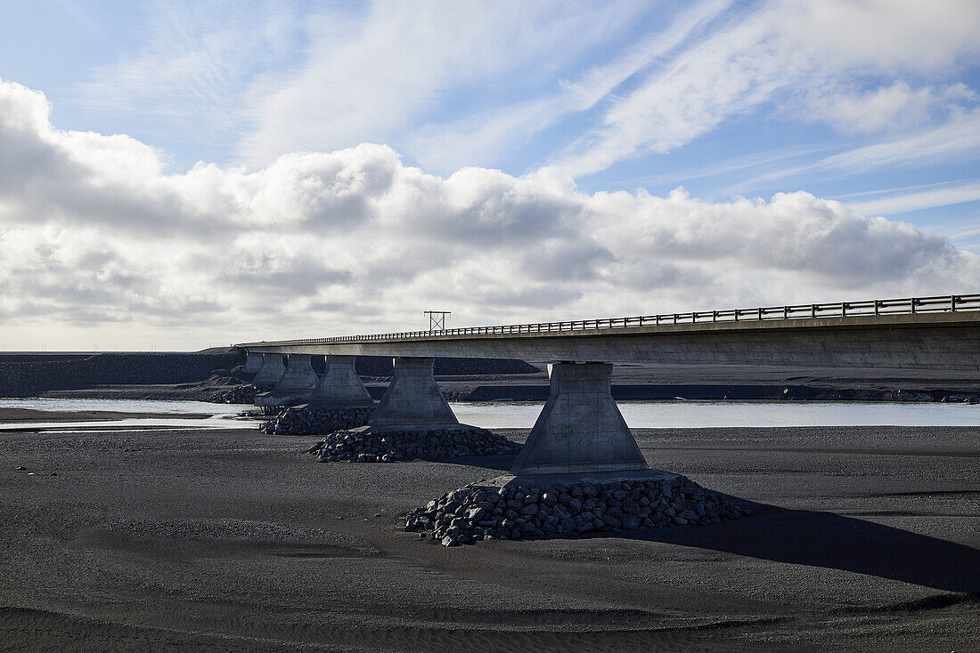 Eine Brücke, die über einen Fluss führt, der durch ein Flussbett aus schwarzem Sand fließt; Island