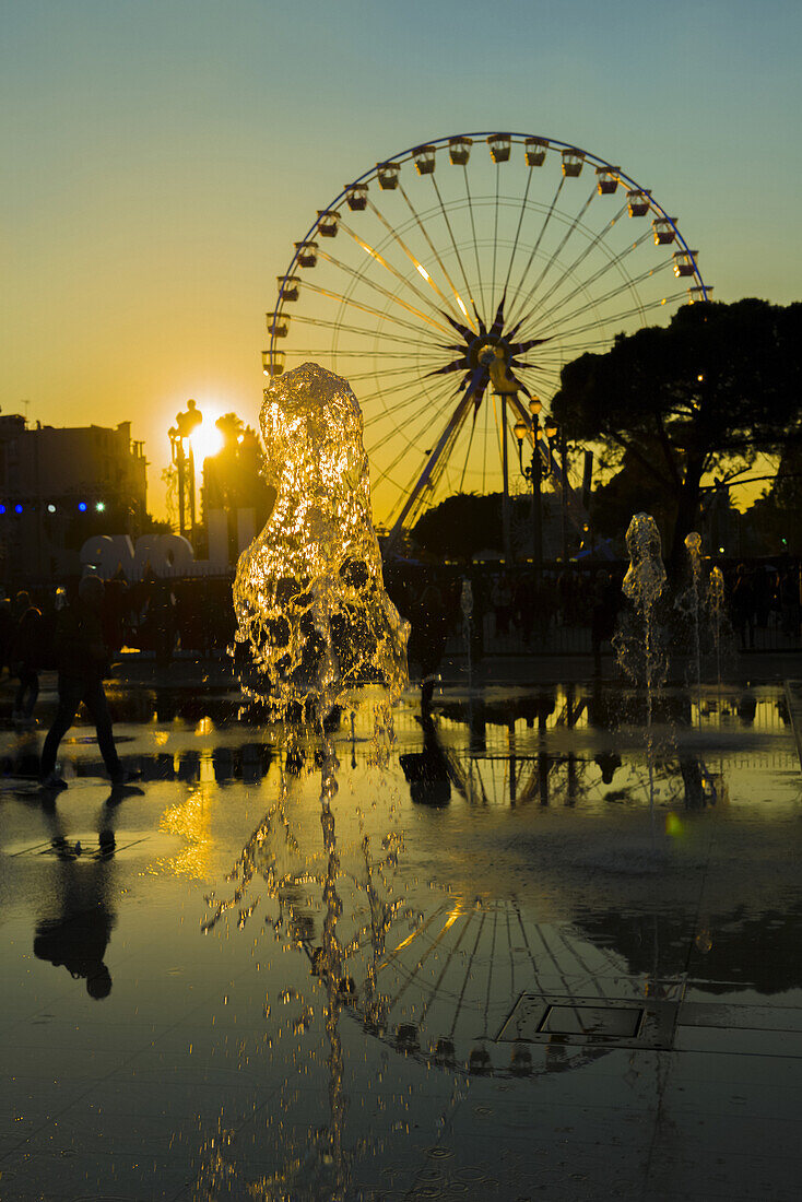 Springbrunnen und ein Riesenrad bei Sonnenuntergang, das sich im Wasser spiegelt, Place Massena; Nizza, Cote D'azur, Frankreich