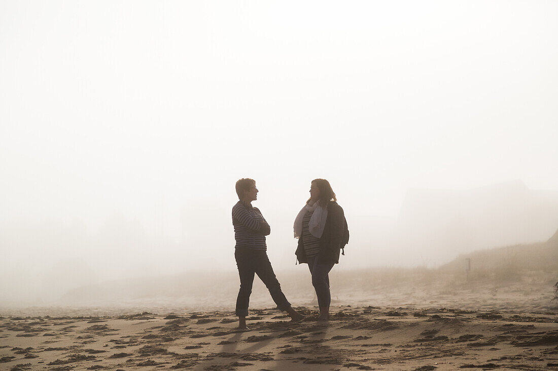 Two Female Friends Stand Talking On A Beach In The Mist; Prince Edward Island, Canada