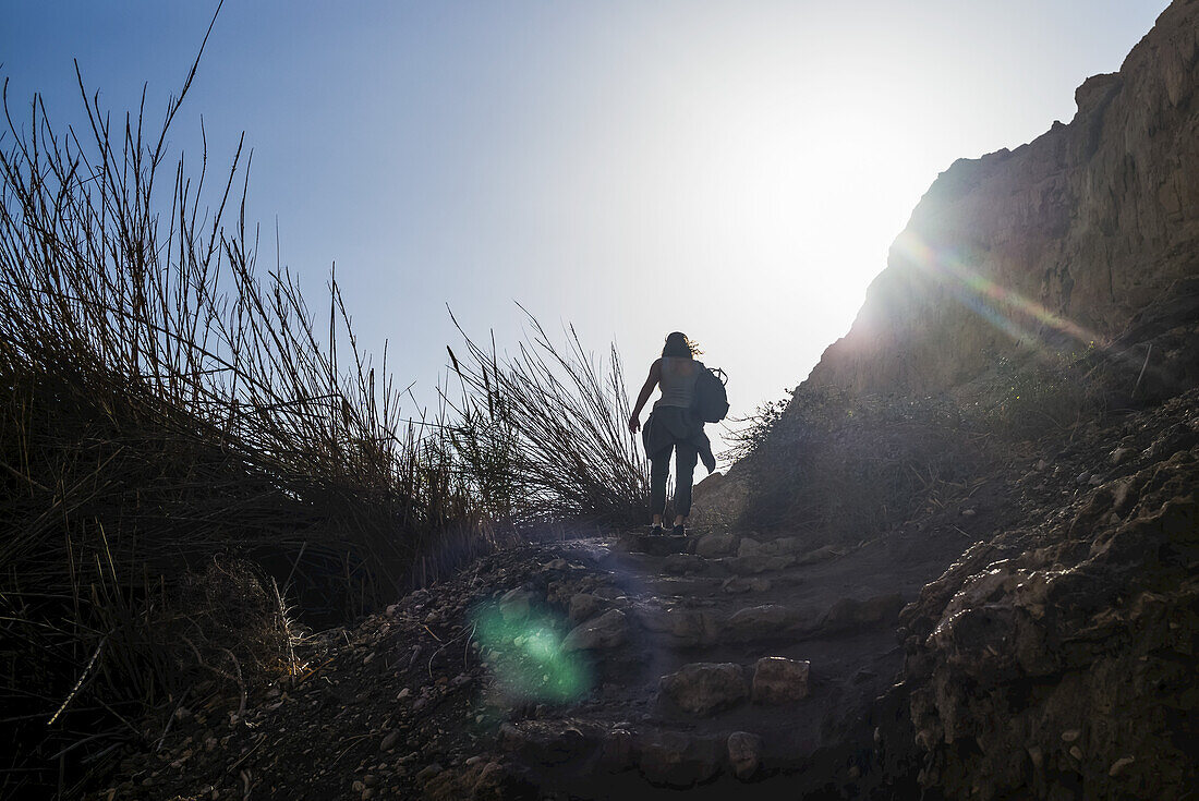 A Young Woman Hikes Up Rugged, Stone Steps In Ein Gedi, With The Sunlight Illuminating The Sky; South District, Israel