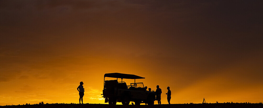 Silhouette von Touristen auf einer Luxussafari, die neben einem Freizeitfahrzeug in der Wüste bei Sonnenuntergang stehen; Sossusvlei, Hardap Region, Namibia.