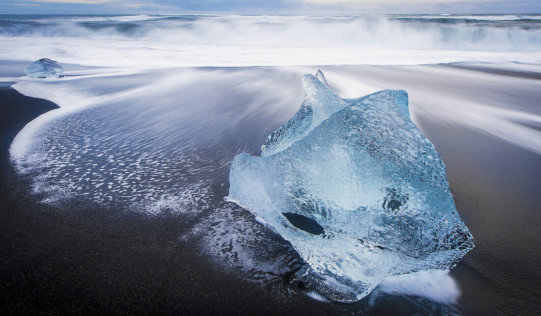 Jagged piece of broken ice laying on the sandy shore at the edge of the ocean water; Iceland