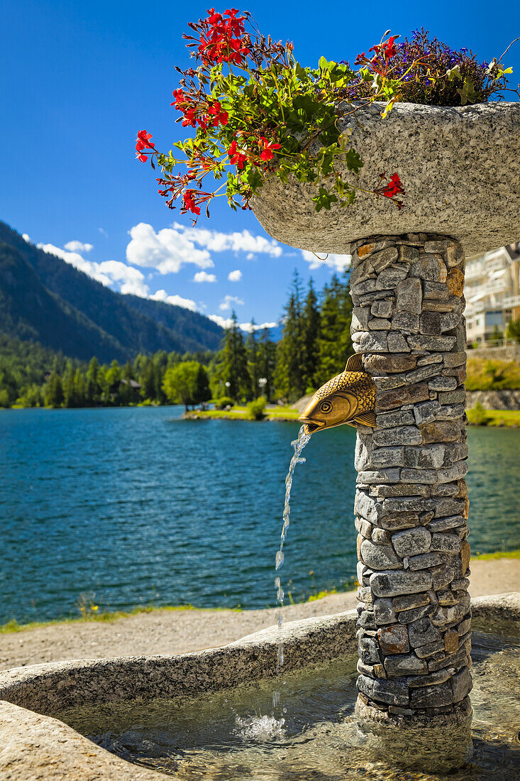 Close-up of a stone fountain with flower pots by Champex Lake and mountain range in the background; Champex, Valais, Switzerland