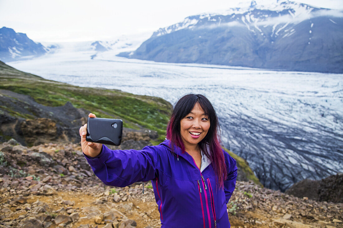 Eine asiatische Wanderin macht ein Selbstporträt auf dem Berggipfel mit dem Gletscher im Hintergrund im Vatnajokull-Nationalpark; Island
