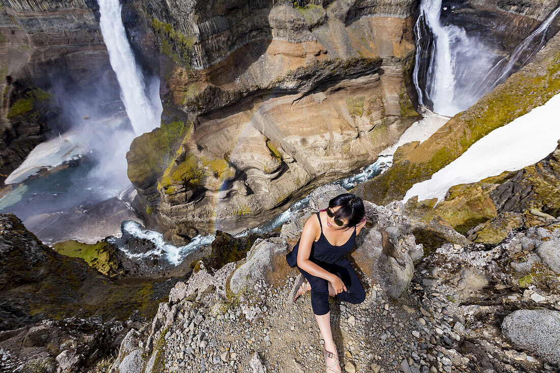 Eine junge asiatische Wanderin posiert für ein Porträt am Rande einer atemberaubenden Landschaft mit zwei Wasserfällen, die als Haifoss bekannt ist; Island