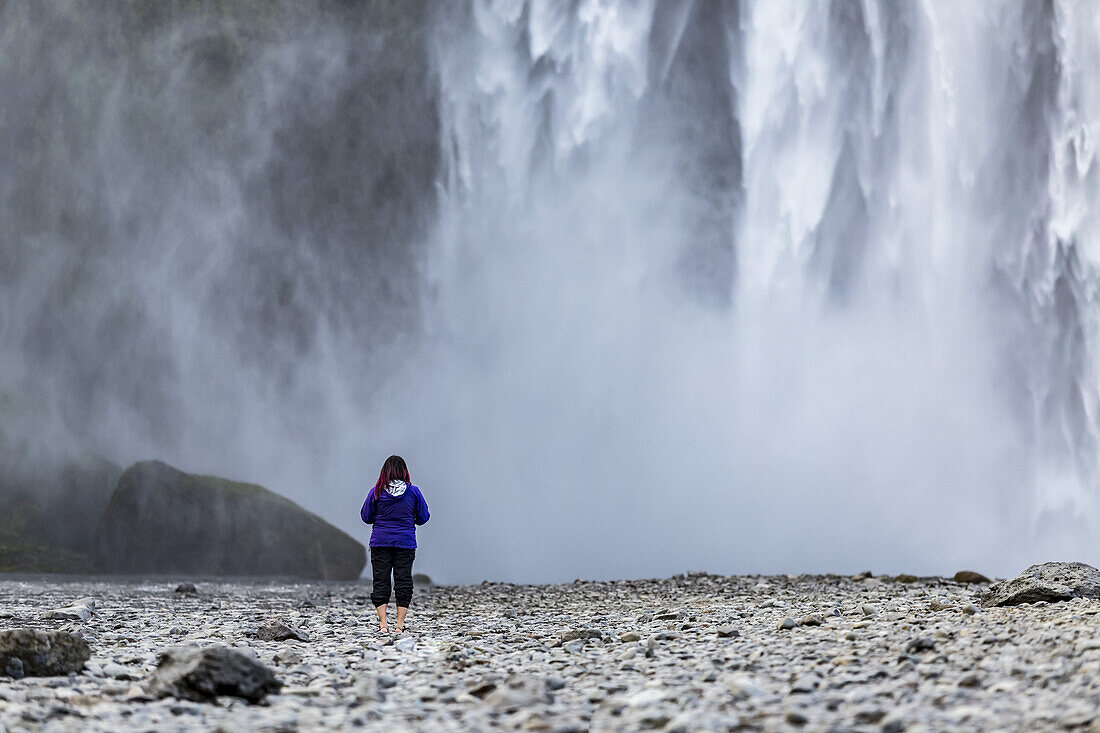 Eine Touristin blickt auf die Wasserwand des berühmten isländischen Wasserfalls Skogafoss; Island