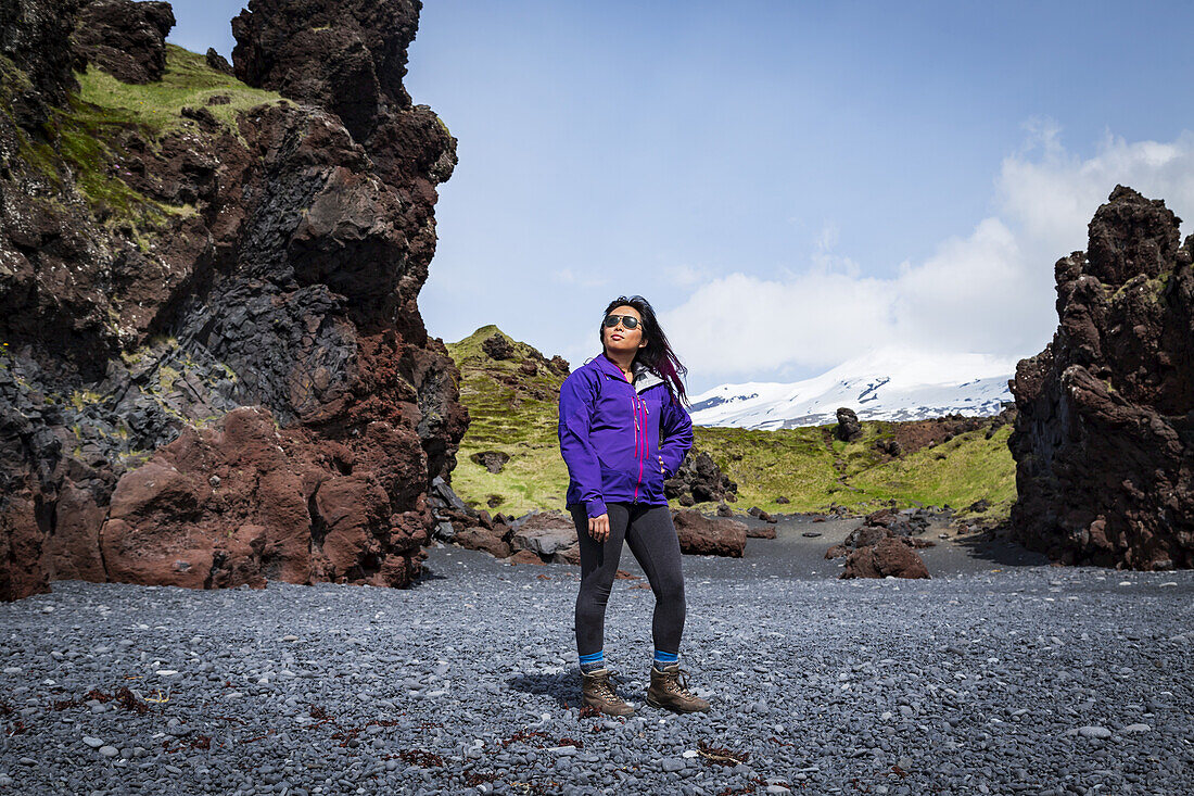 An Asian female tourist poses for a portrait on the black sand beach next to a rock formation in Western Iceland, Snaefellsnes peninsula; Iceland